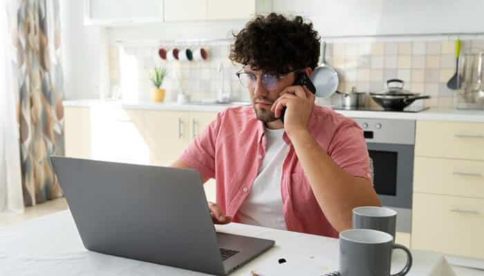 A person working from home, sitting at a kitchen table with a laptop and talking on the phone, maintaining workplace culture remotely