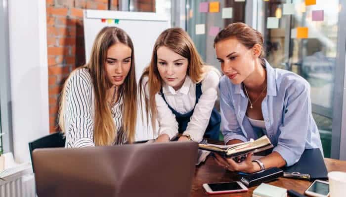 Three young female entrepreneurs collaborating at a workspace, studying smart and planning their business strategy using a laptop and notes