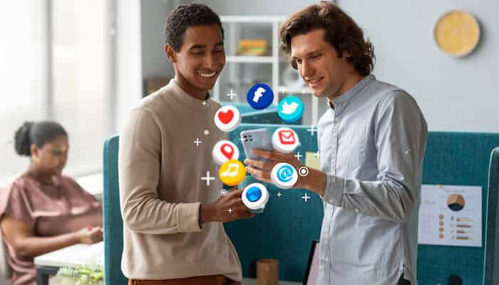 Two young men discussing Social Media Marketing with floating icons of Facebook, Twitter, Instagram, LinkedIn, and other platforms in a modern office setting, while a woman works in the background