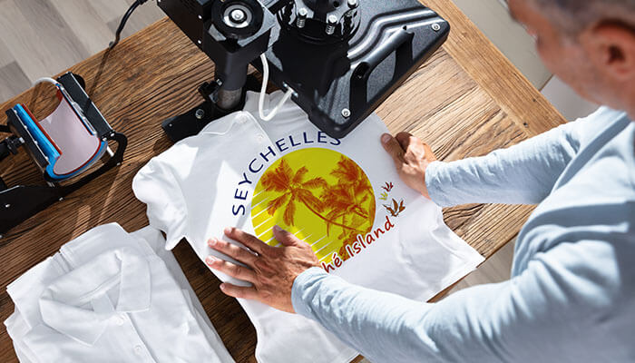 trends in t-shirt printing person heat-pressing a colorful Seychelles palm design onto a white T-shirt on a wooden table