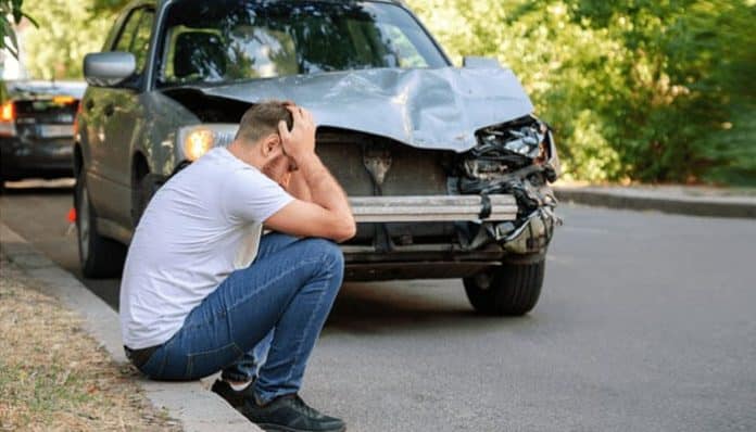 Person sitting on the roadside in distress after a car accident, with front-end damage to the vehicle, reflecting the stress of being partially at-fault for an accident.