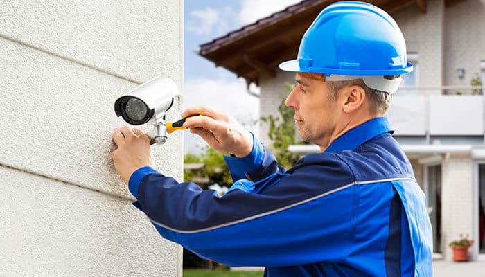 A professional technician wearing a blue uniform and safety helmet installing a CCTV camera on a building wall, representing reliable installation and CCTV home security solutions for enhanced safety.