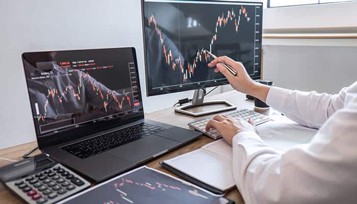 A trader analyzing stock market charts on dual monitors and a laptop, demonstrating technical analysis and decision-making strategies in swing trading.