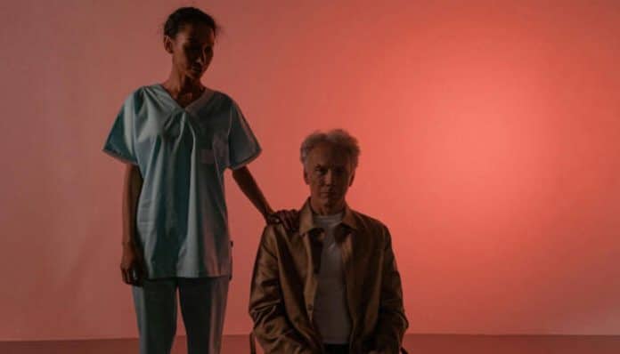 A caregiver standing beside an elderly resident in a dimly lit room, representing the skills and support taught through assisted living facility training programs.