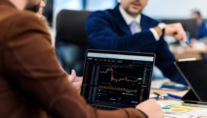 Two professionals discussing strategies while viewing a prop trading chart on a laptop, highlighting the process of launching and managing a prop trading firm