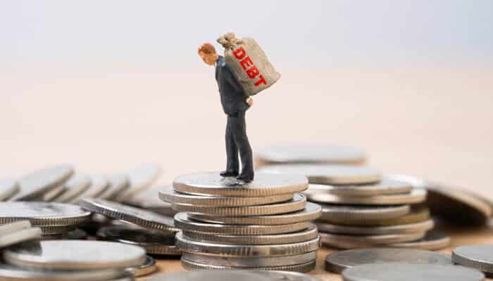 Miniature businessman standing on coins carrying a heavy debt bag, visually representing the psychology of consumer debt traps and financial pressure.